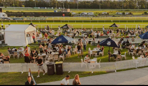A packed lawn of race-goers at tables and umbrellas beside the racetrack